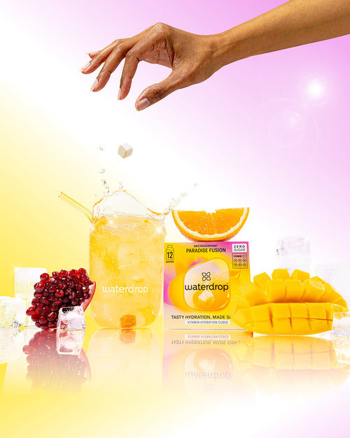 Hand dropping Waterdrop's PARADISE FUSION cube into fizzy glass, surrounded by pomegranate, mango, orange, and ice, with 12-serving pack nearby, highlighting a tropical refreshment.