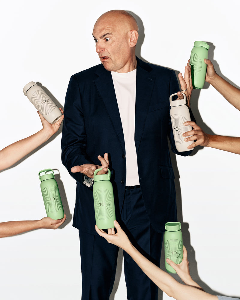 Man wearing a dark suit stands as multiple hands present assorted green and white water bottles around him.