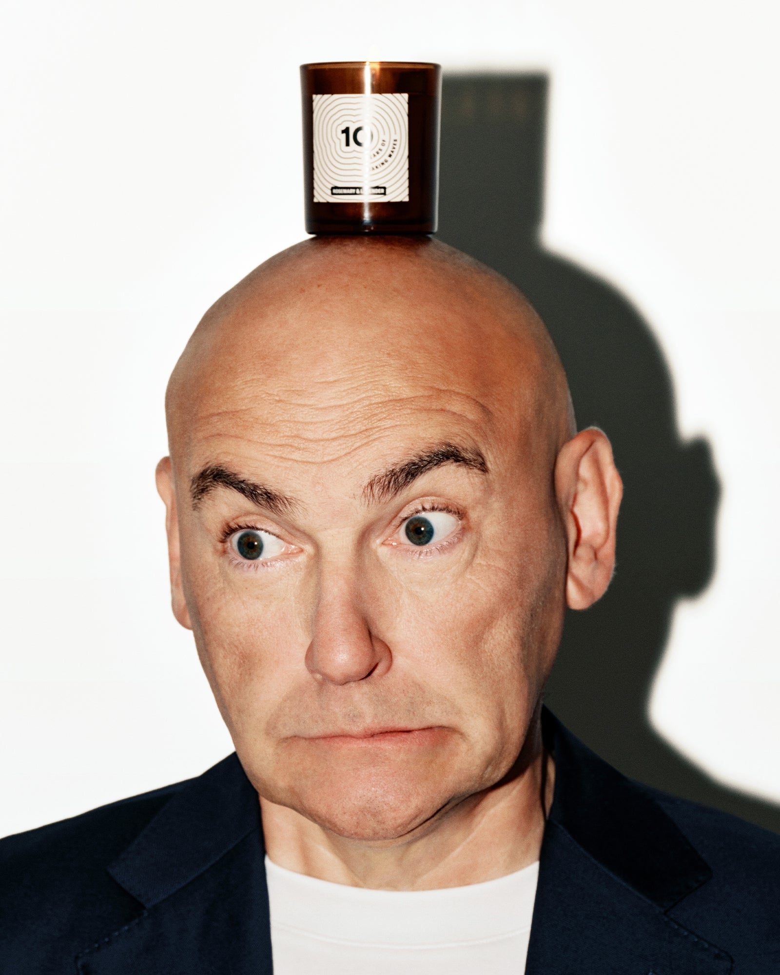 Bald man wearing a dark blazer with a small copper bottle balanced on his head.