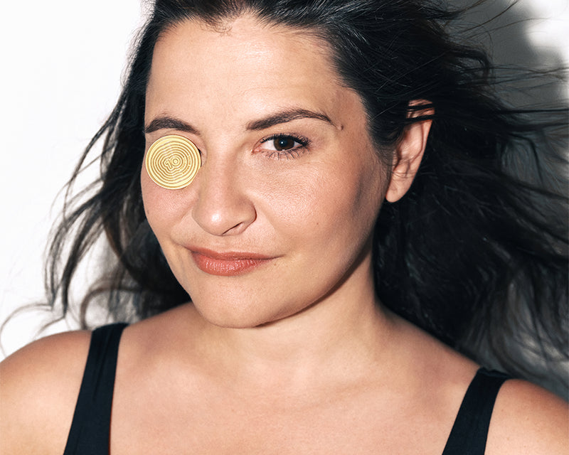 Woman with dark hair wearing a black tank top, facing the camera, with a gold circular patch on her left cheek near the eye.