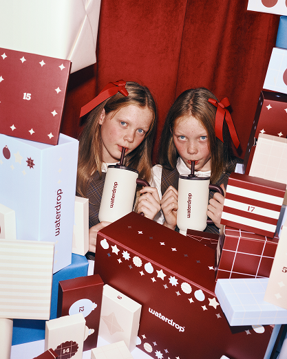 Two girls drink from white cups among colorful wrapped gifts.