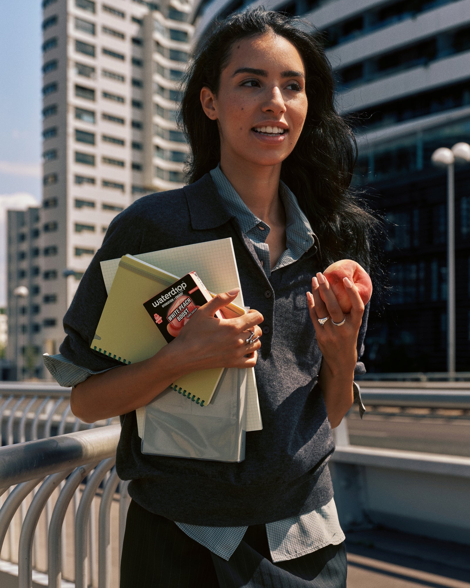 A woman walks outdoors in an urban setting, carrying notebooks and a card.
