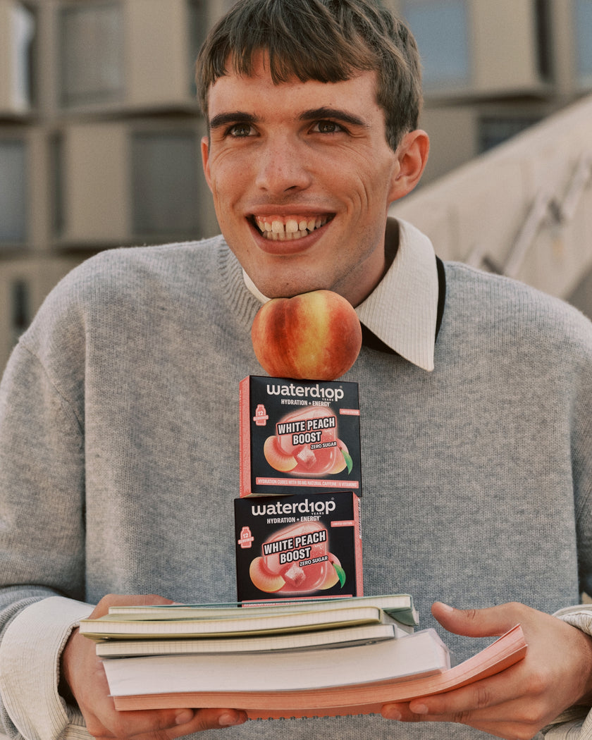 Young man in a gray jumper holding two peach-flavored boxes on top of a stack of books.