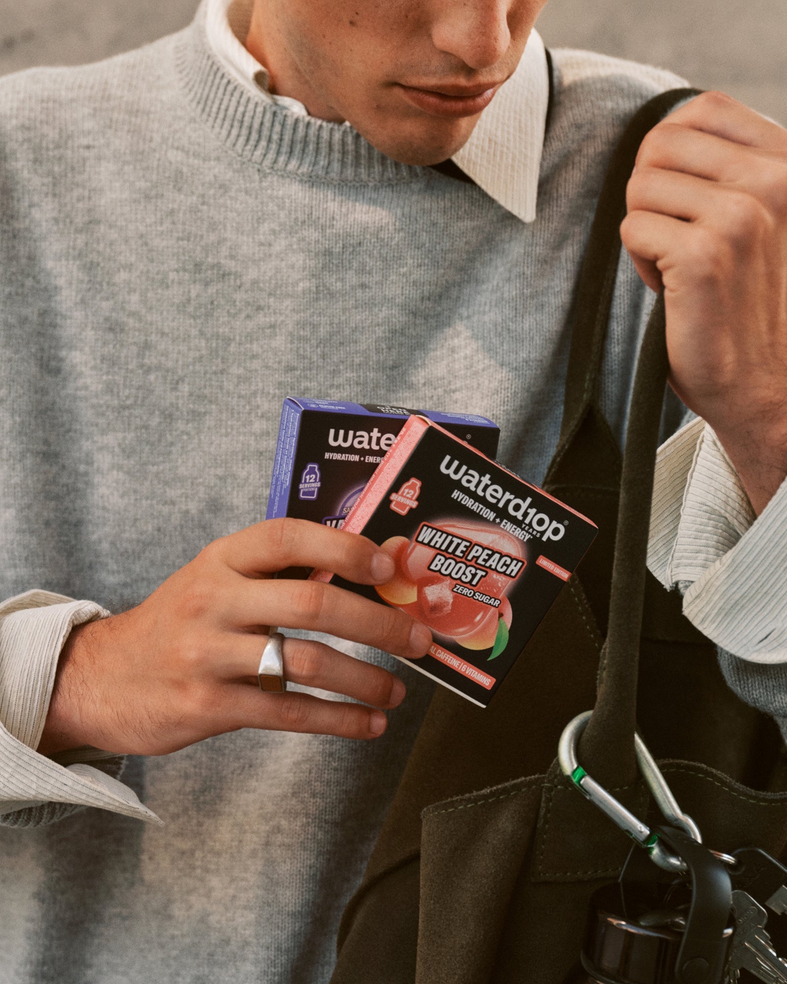 Person in a gray sweater holding small product boxes beside a black bag.