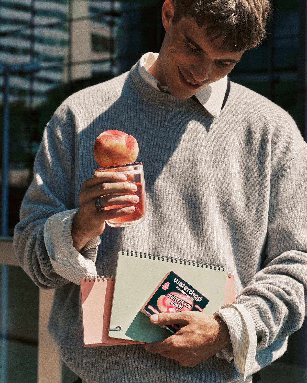 Man in a gray sweater outdoors holds a pink beverage with an apple on the rim and a notebook in his other hand.