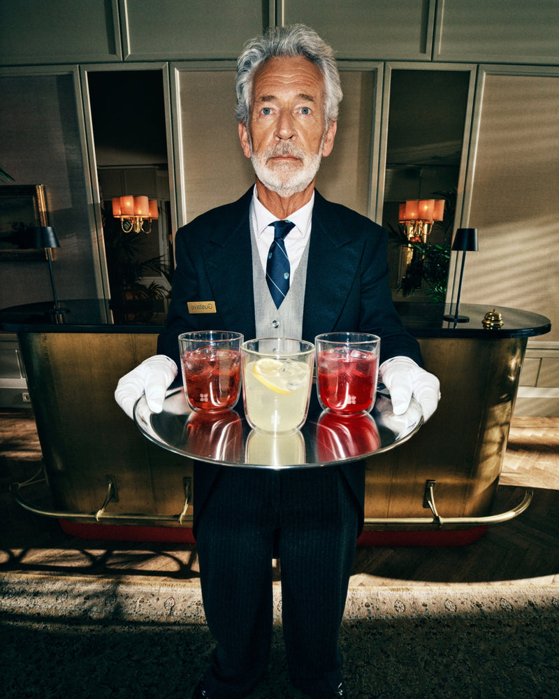 Senior waiter in a dark suit presents a round tray with two red drinks and one pale-yellow drink in a hotel lobby.