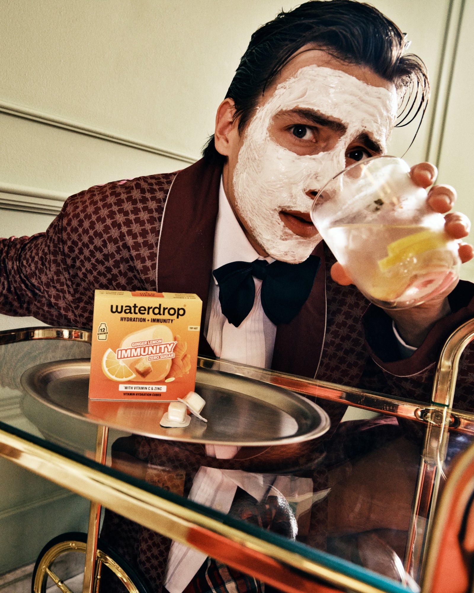 Man in a brown suit wearing a facial sheet mask and drinking from a glass at a bar.