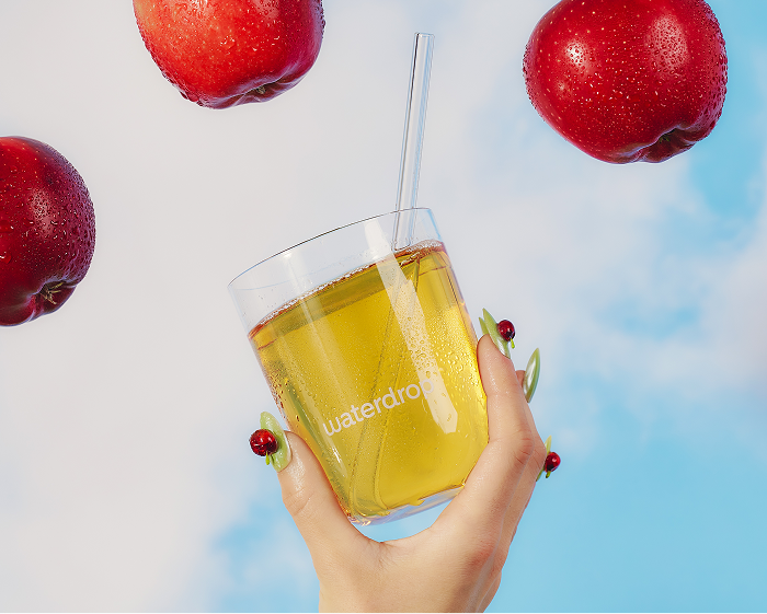 Hand holding a glass of yellow juice with a straw, surrounded by red apples against a blue sky.