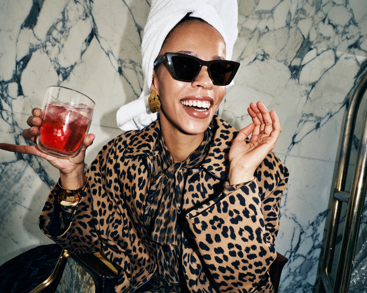 Woman wearing a leopard-print coat and sunglasses, with a white towel on her head, holding a red beverage in a glass against a marble wall.