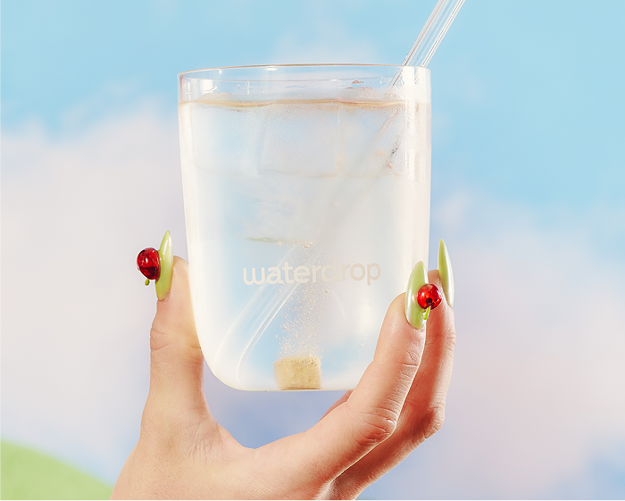 A hand holds a clear glass of water with ice and a straw against a blue sky.