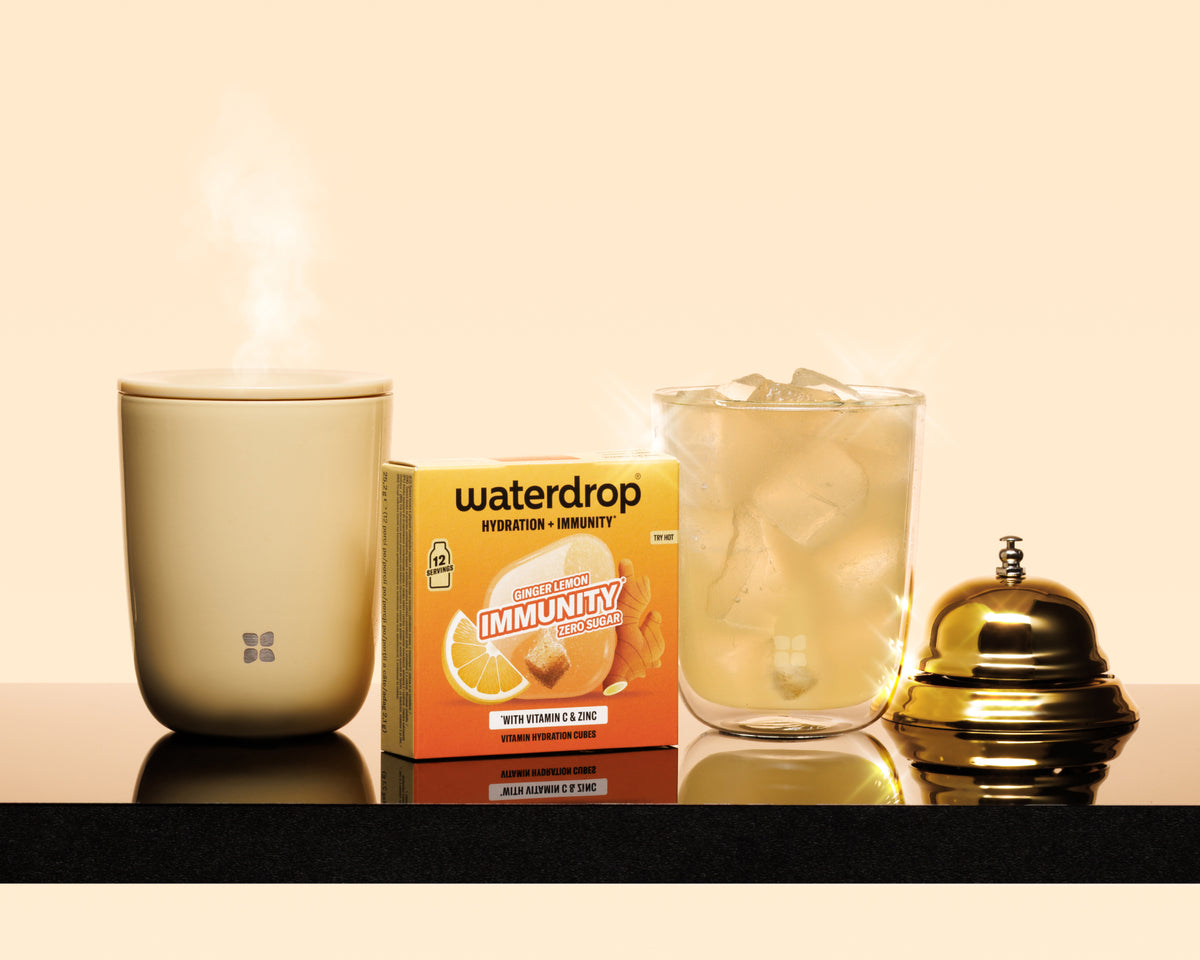 Steaming beige mug, boxed immunity product, glass with ice and a polished gold bell on a black countertop.