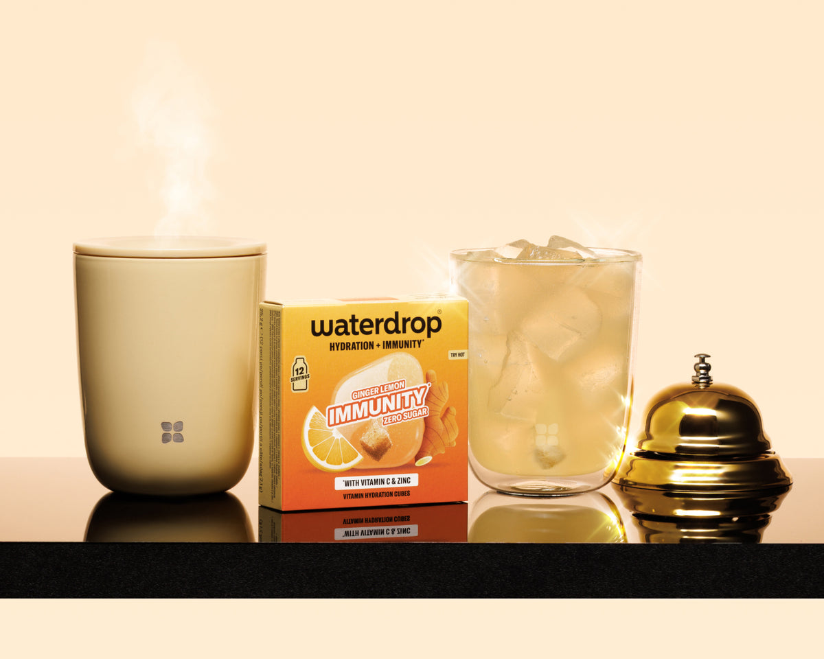 Steaming beige mug, a boxed product, a glass with ice, and a brass service bell on a dark shelf.