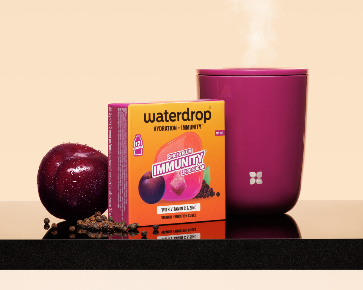 Purple cup with steam beside an orange Waterdrop box, a plum and scattered seeds on a reflective surface.