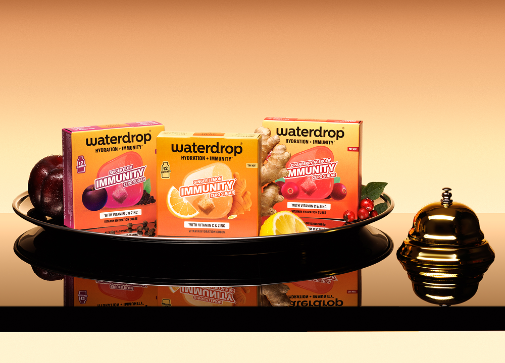 Assorted drink-mix boxes displayed on a black tray with a reflective surface and a small gold bell on the right.