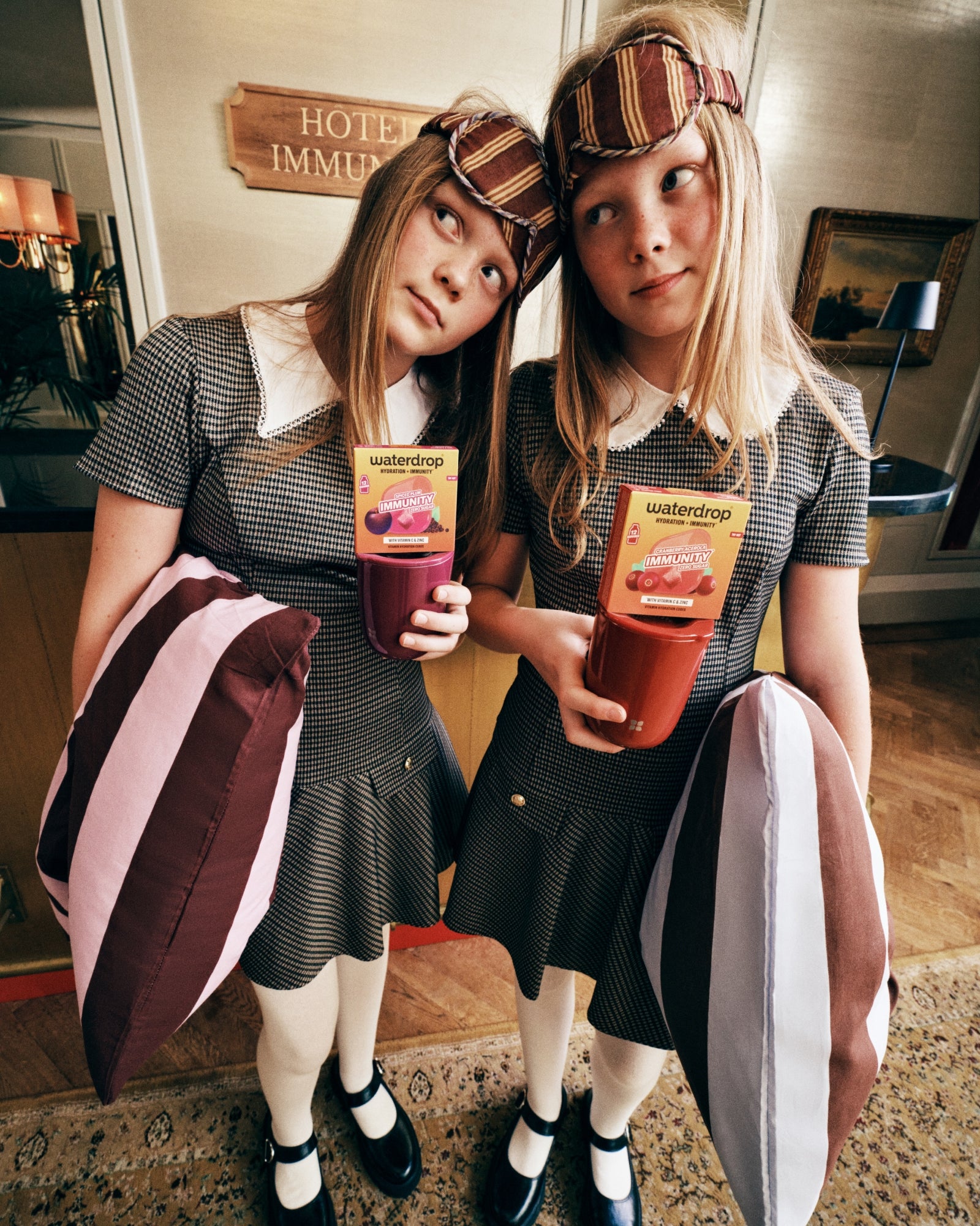 Two girls in matching checkered dresses with striped headbands stand side by side in a hotel lobby, each holding a juice pouch.