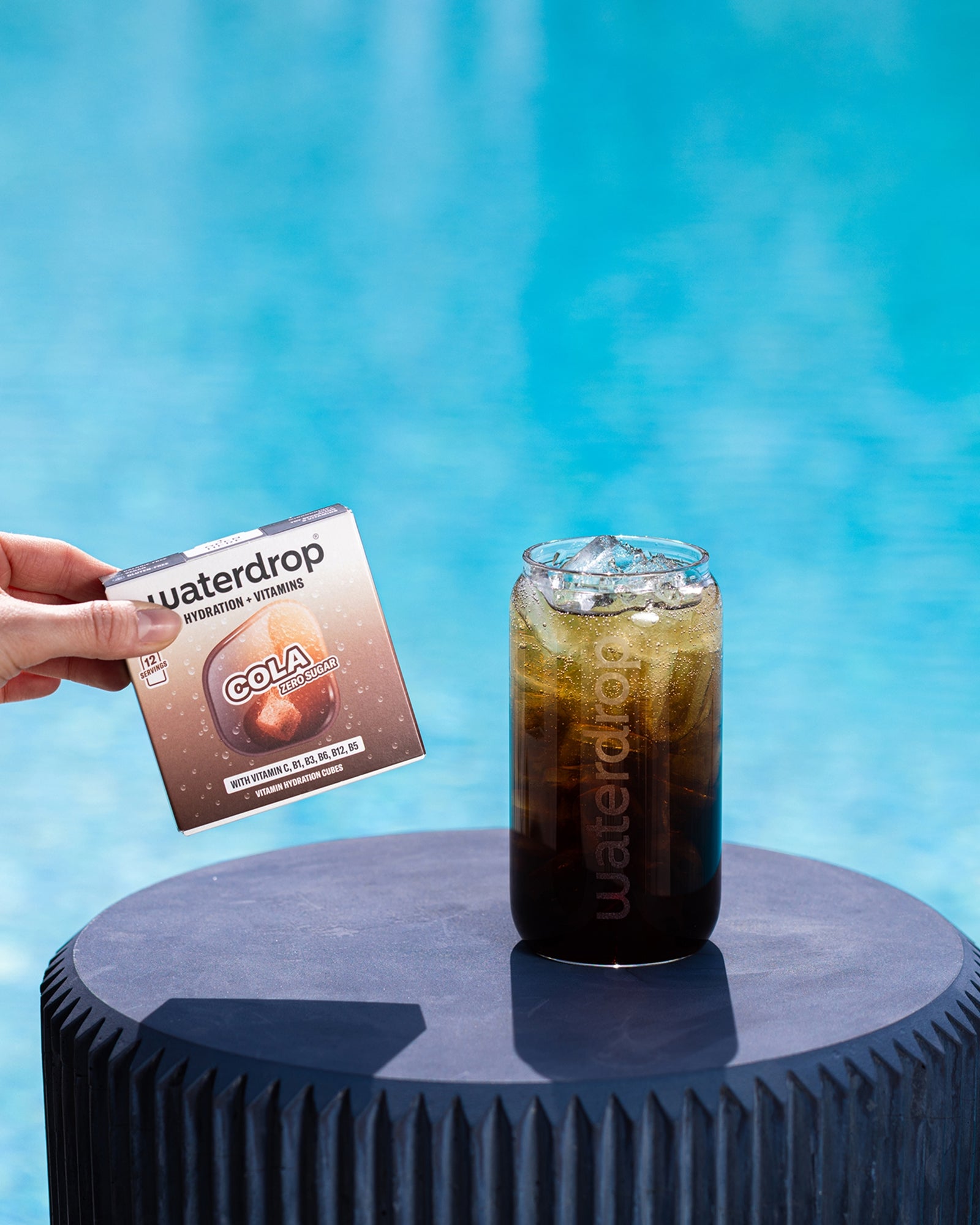 Hand holds a small packet beside a glass of iced cola on a round blue pedestal by a pool.